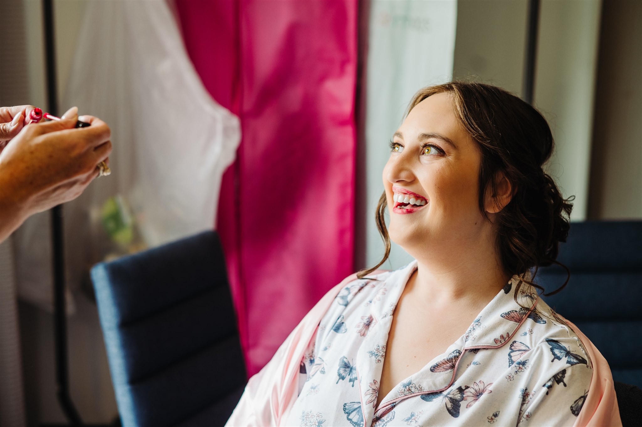 Woman in floral robe smiling while red lipstick is applied, pink and white curtains in background.