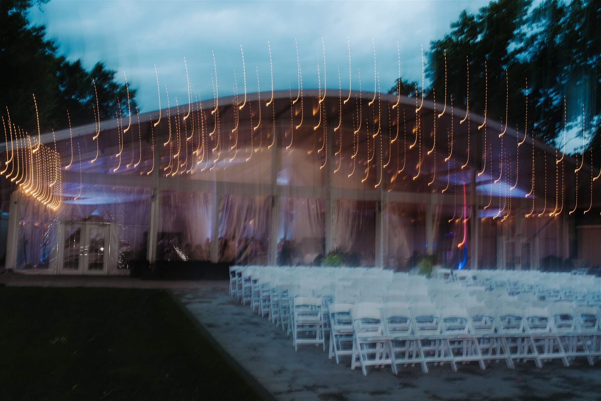 Outdoor evening event setup with rows of chairs facing a tent decorated with string lights and sheer drapes.