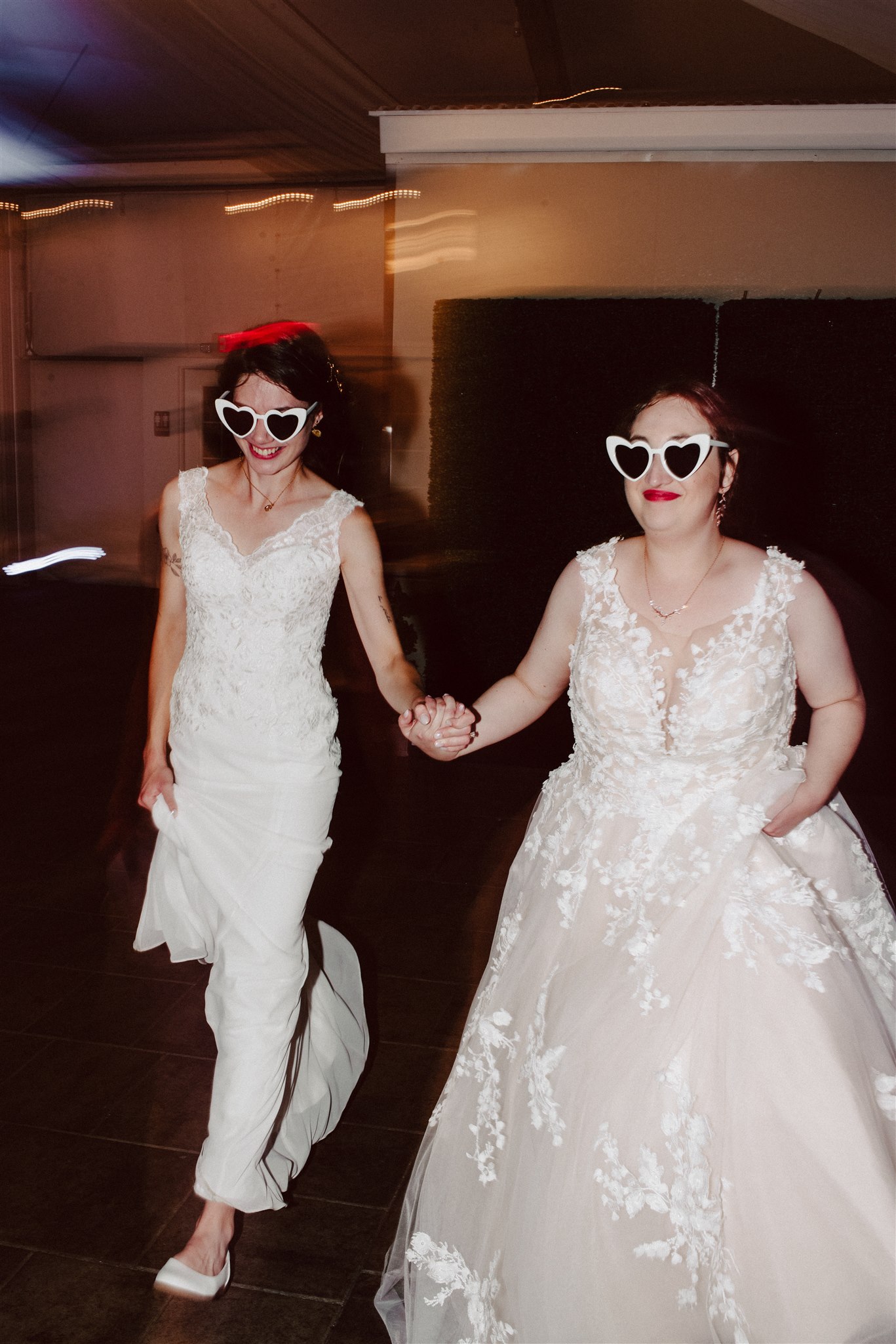 Two people in ornate white dresses and heart-shaped sunglasses walk hand-in-hand in a dim setting.