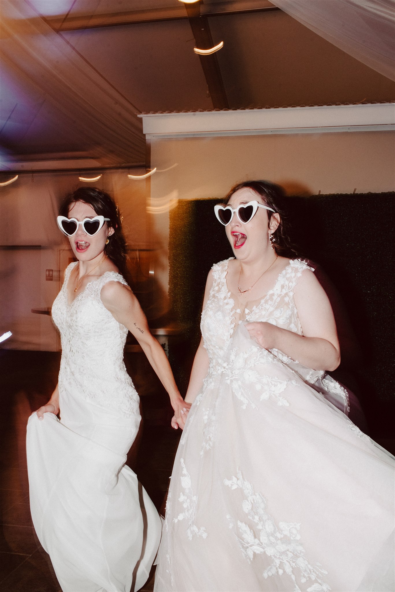 Two brides in lace wedding gowns wearing heart-shaped sunglasses dance and laugh joyfully indoors.