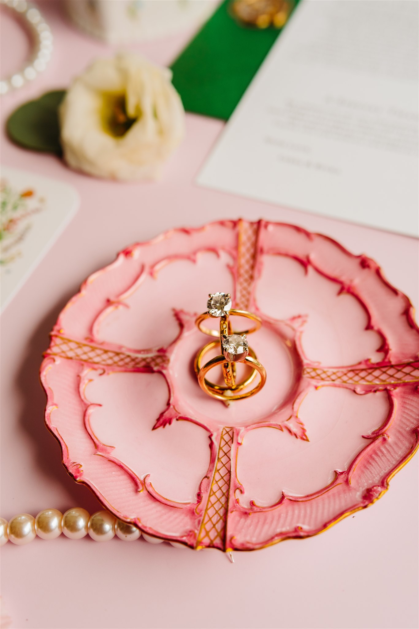 Two diamond rings on a decorative pink plate with pearls, white flower, and papers in the background.