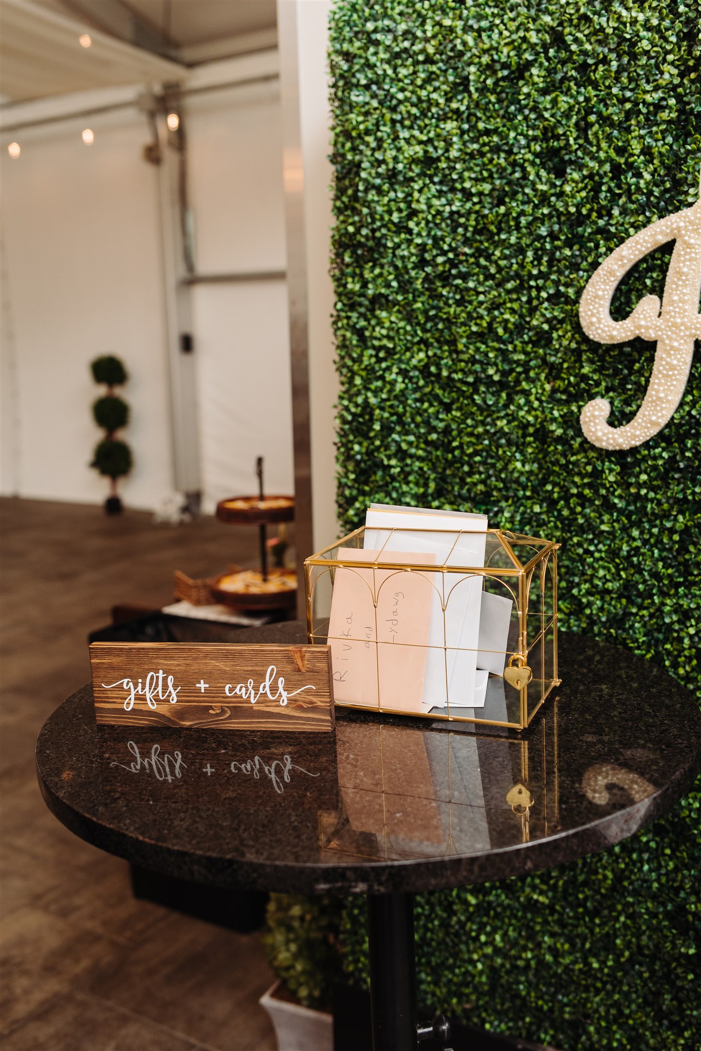 Small black table with a glass card box filled with envelopes and a "gifts + cards" sign, leafy backdrop.