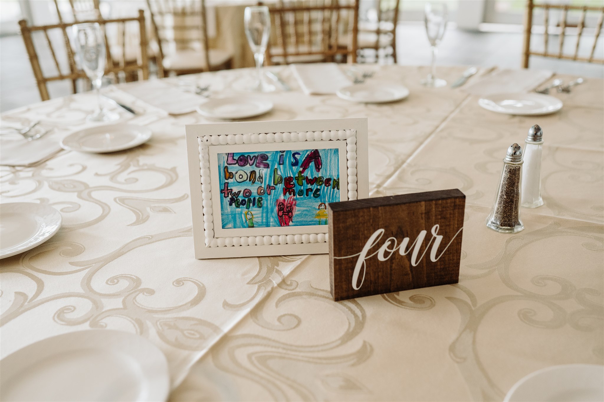 Table set for formal event with framed child's drawing, "four" sign, plates, utensils, glassware.