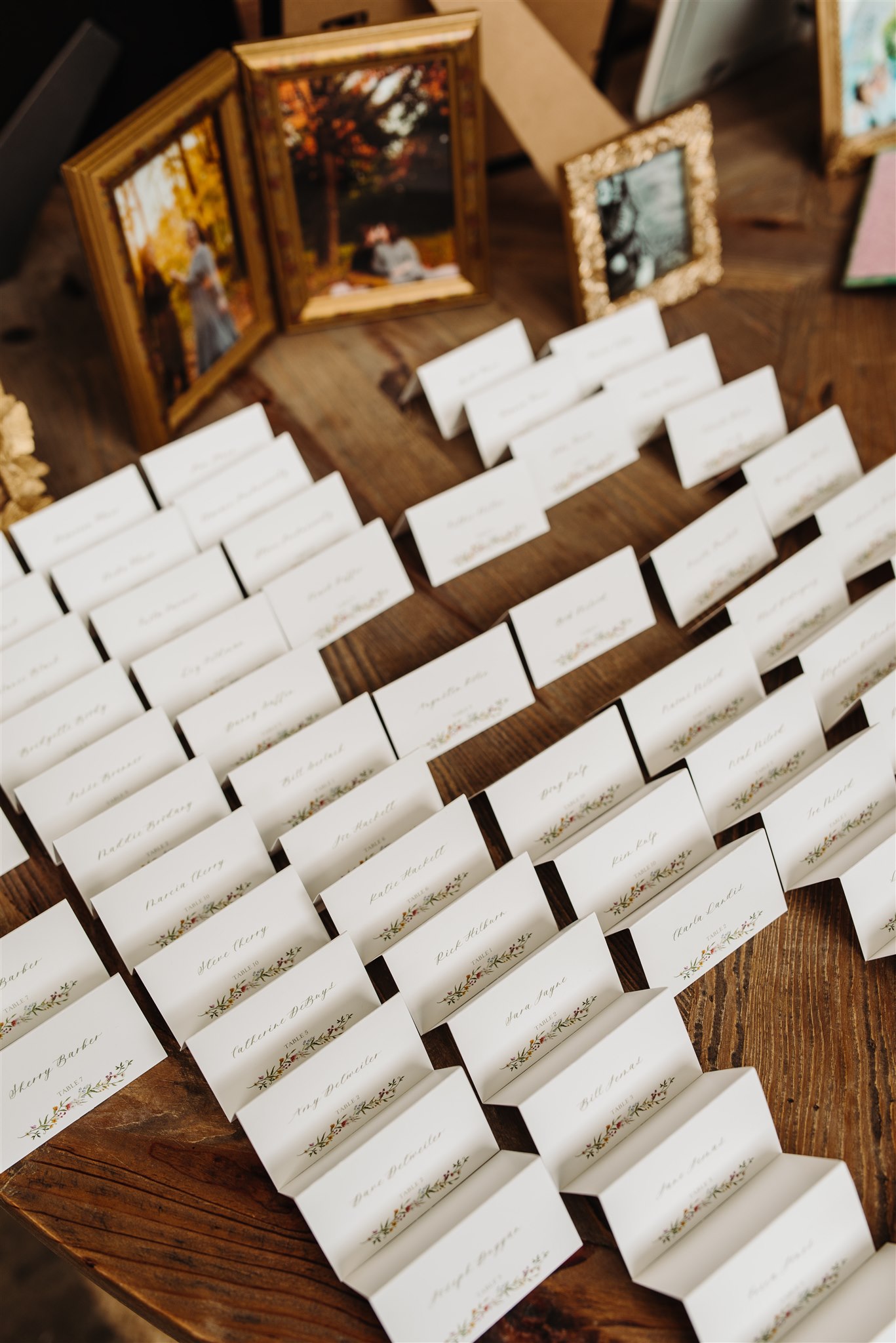 Name cards with floral decorations arranged on a wooden table with framed outdoor photos behind.