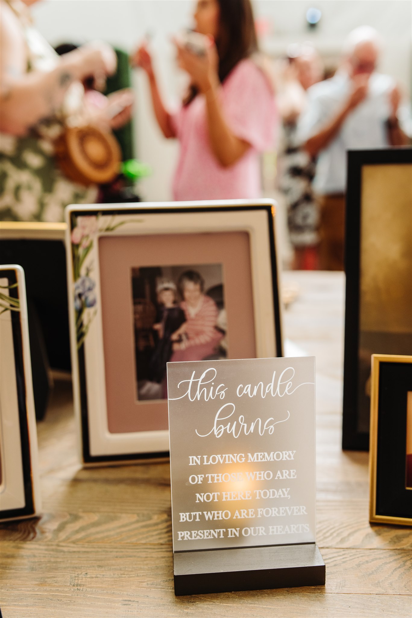 A memorial setup with a framed photo and an acrylic sign honoring absent loved ones, people in background.