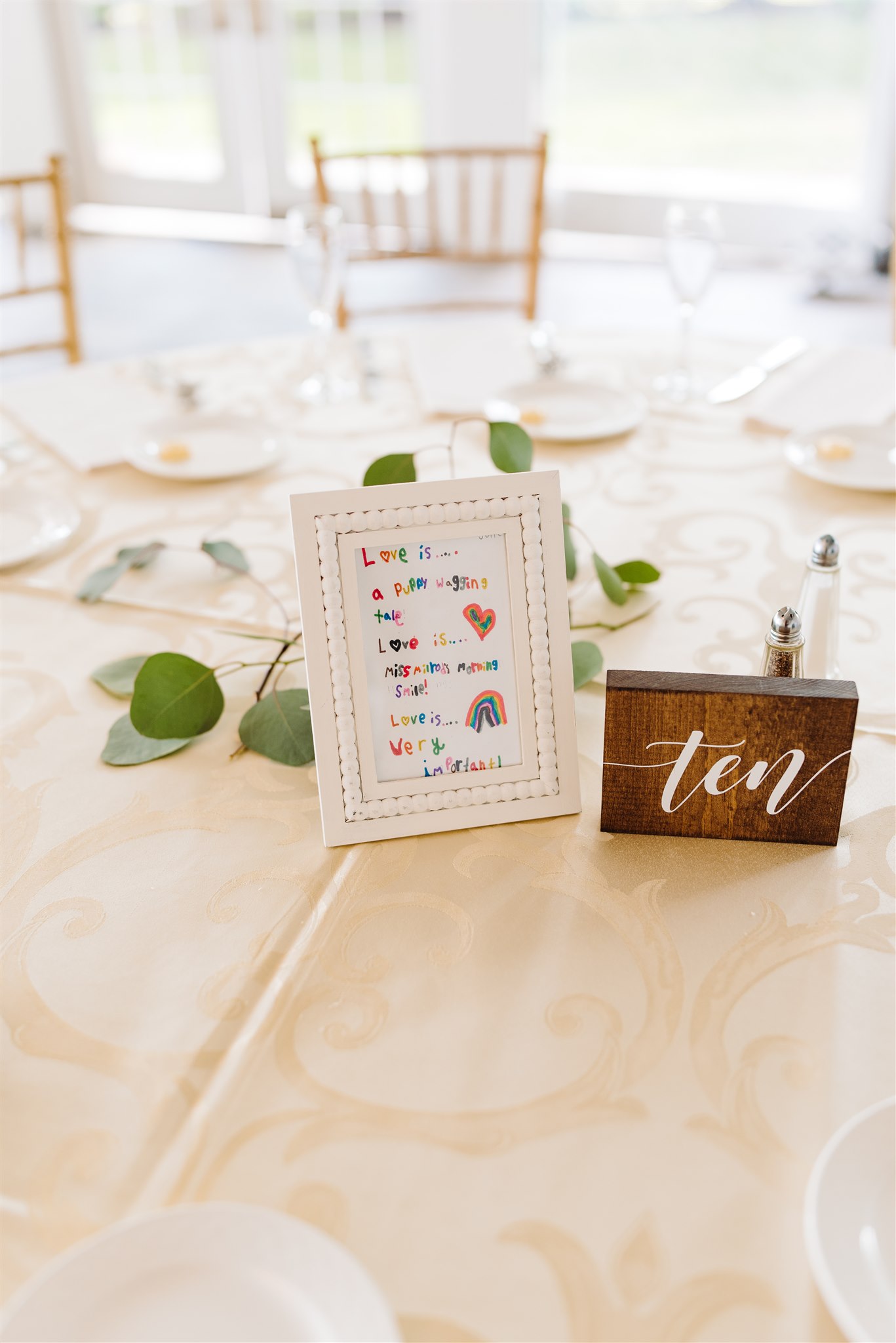 Table with beige tablecloth, framed colorful sign, wooden table number ten, chairs, and greenery.