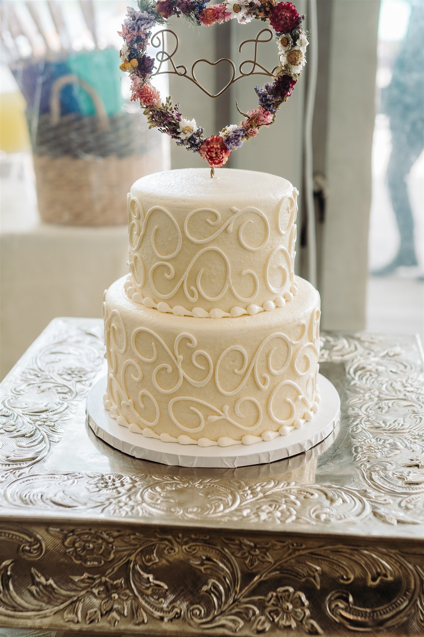 Two-tier white cake with intricate icing patterns on ornate metallic stand, topped with floral heart.