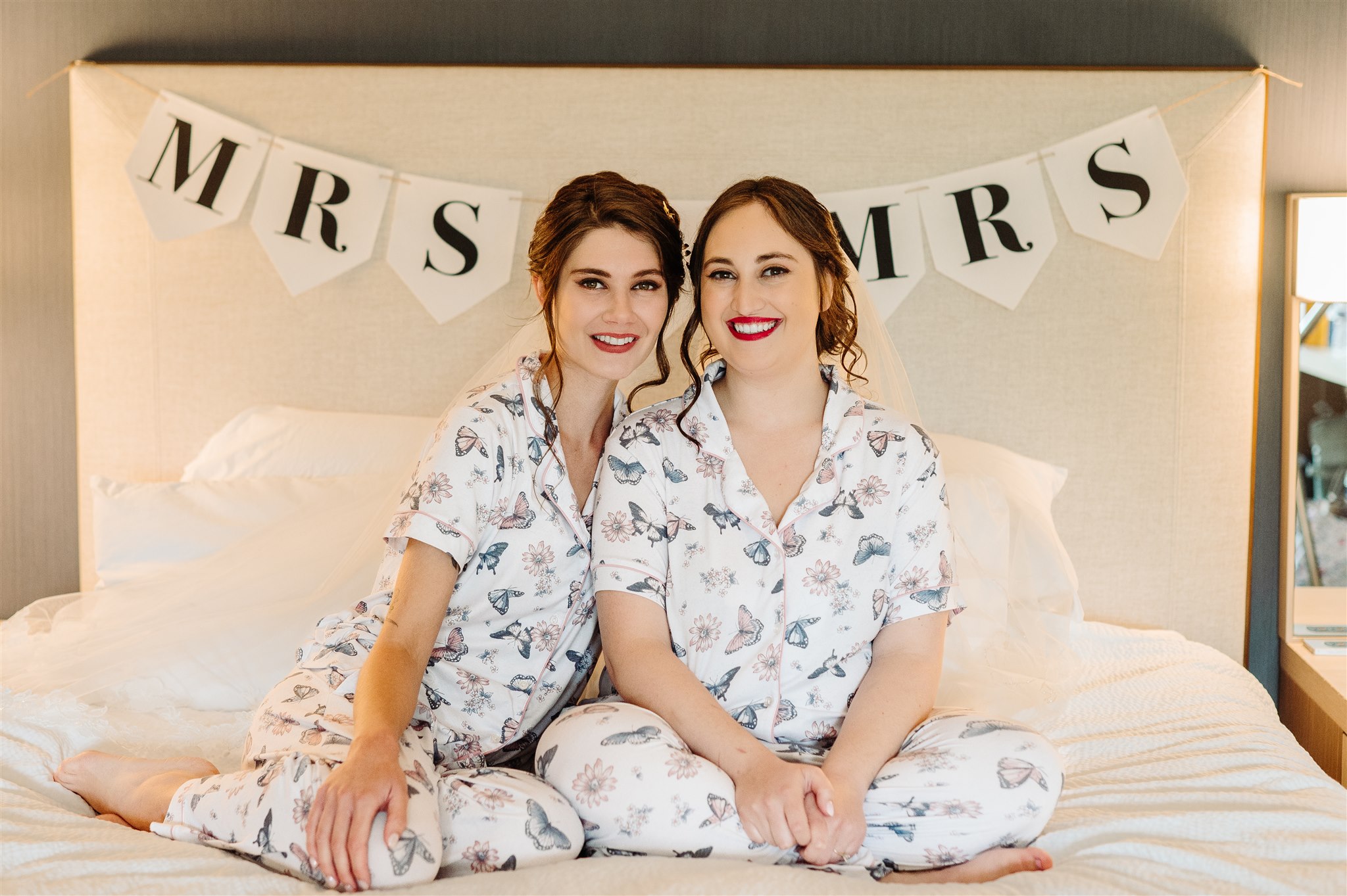 Two people in matching floral pajamas sitting on a bed with a "MRS MRS" banner in the background.