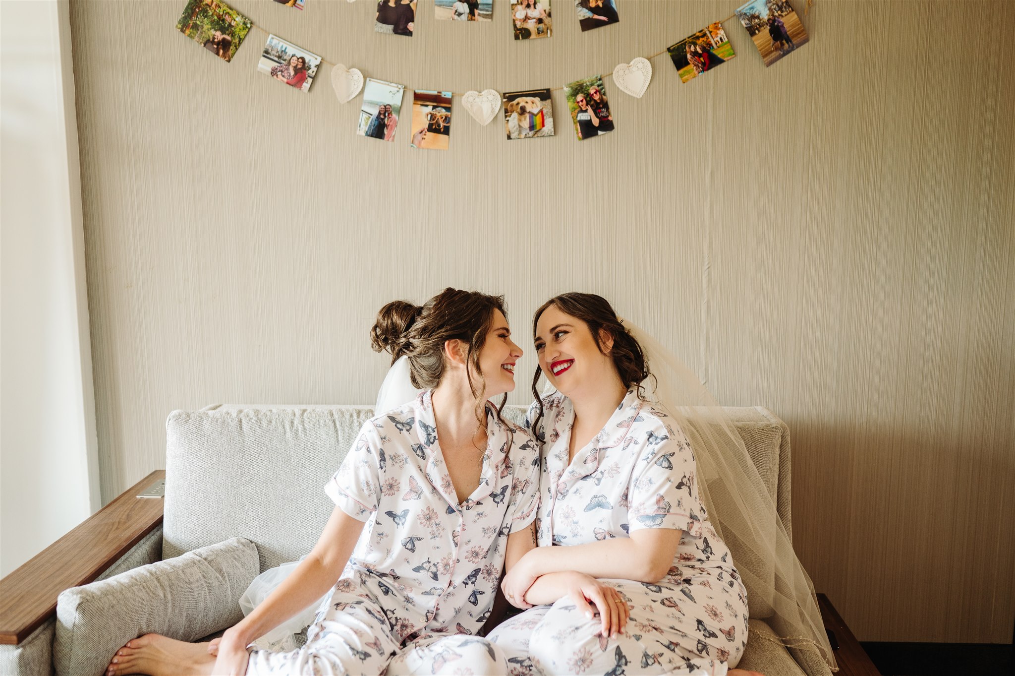 Two women in floral pajamas sit on a couch, with hanging photos and heart decorations above them.