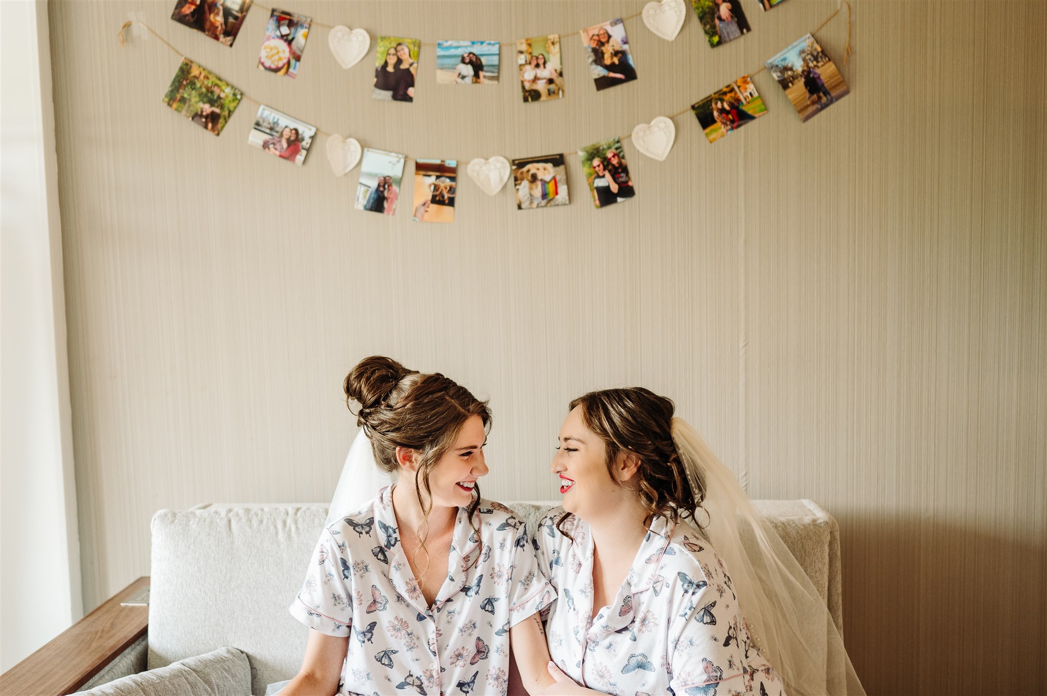 Two women wearing matching floral pajamas and veils sit on a couch, smiling, with photos above them.