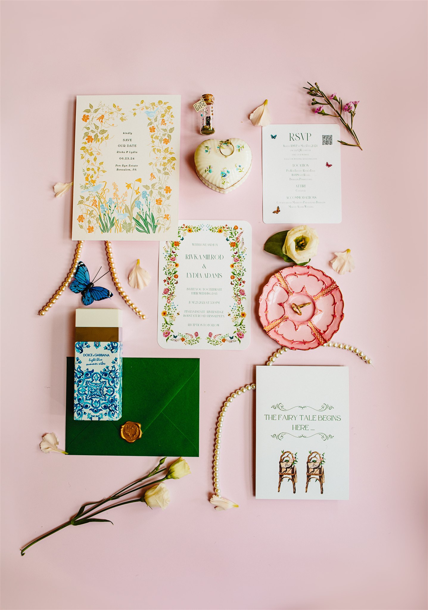 Wedding stationery and accessories on pink background with floral designs, rings, perfume, and envelope.