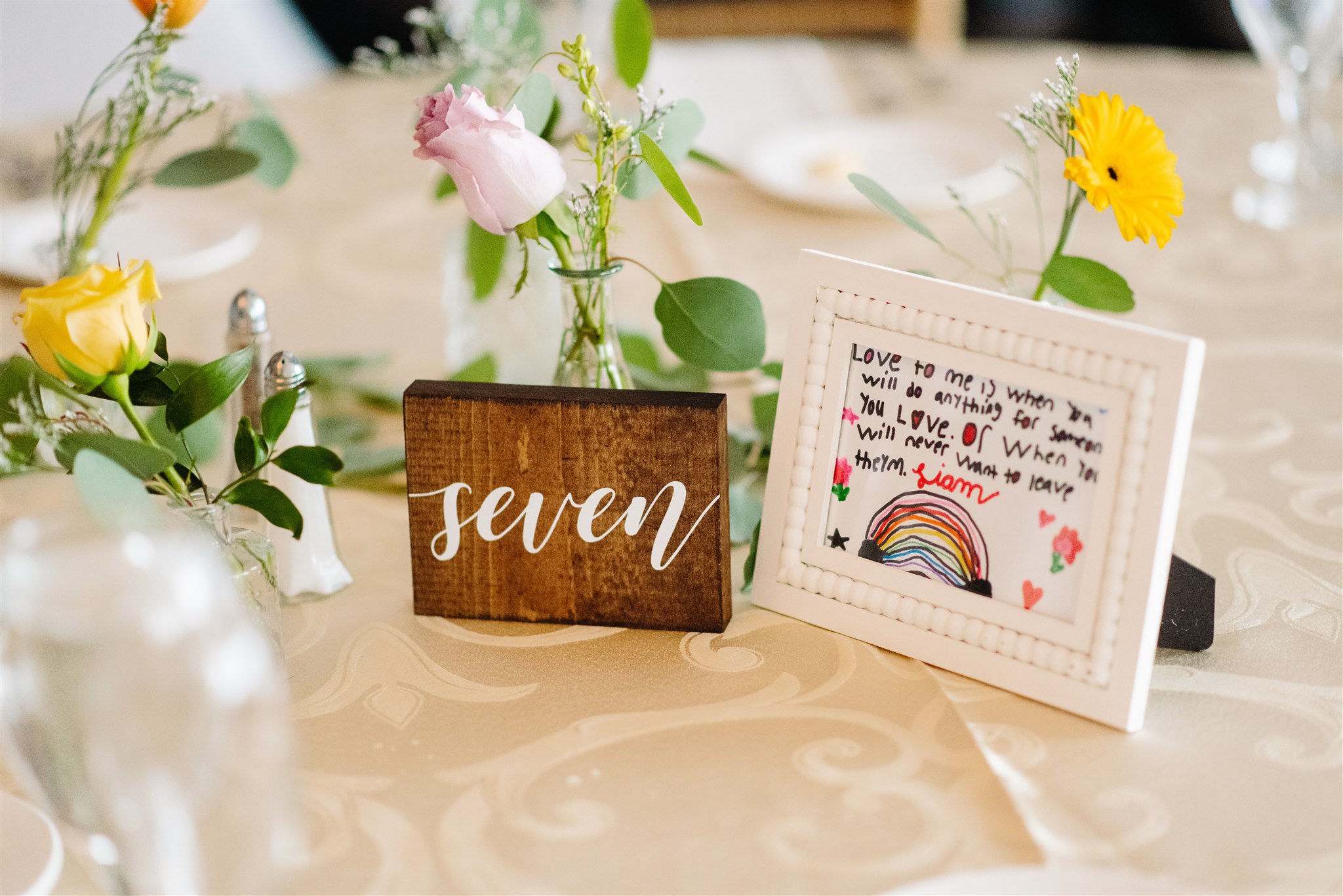 Table with floral arrangements, a wooden "seven" sign, and framed rainbow artwork on a cream cloth.