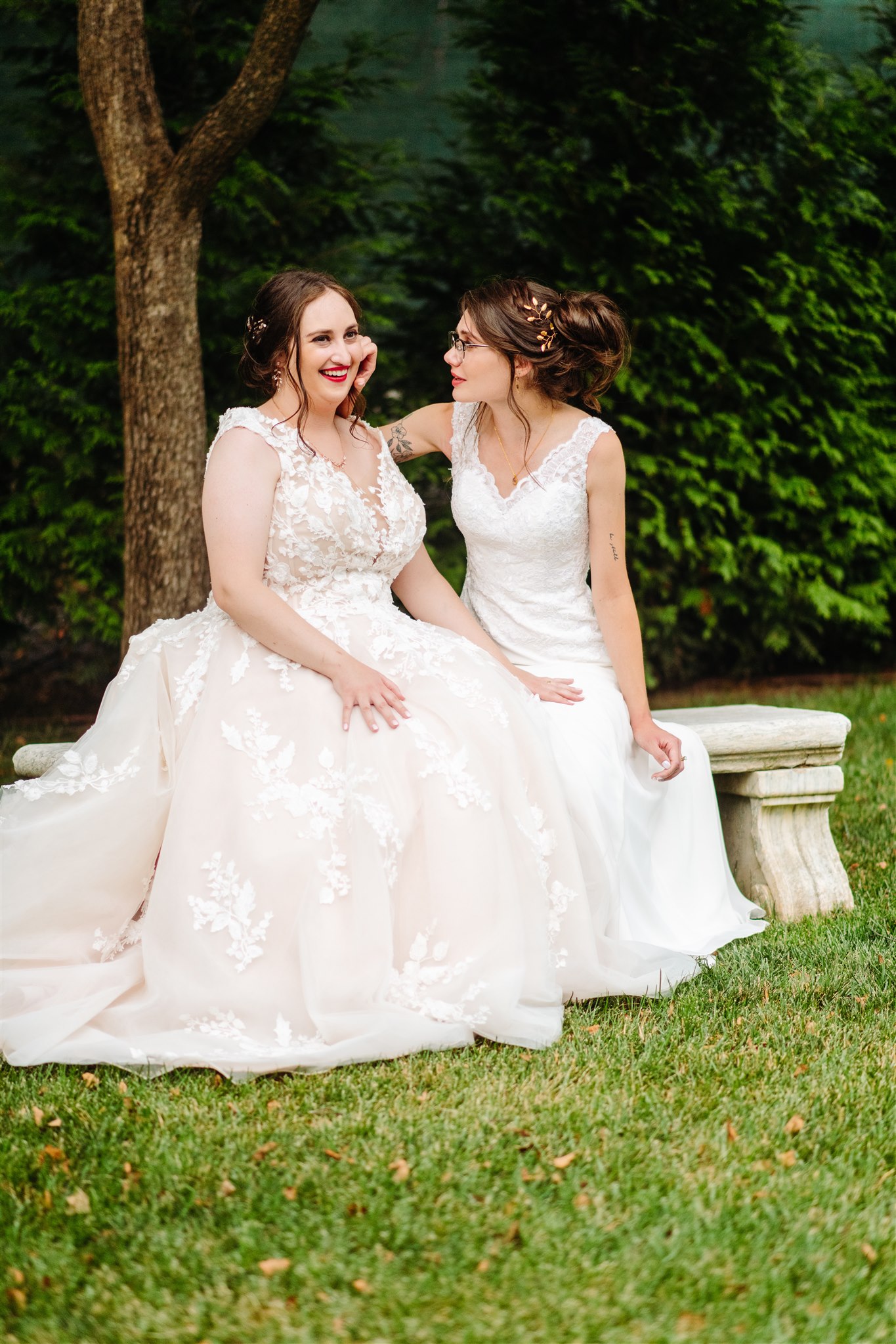 Two women in wedding dresses sitting on a stone bench in a garden, smiling and touching faces.