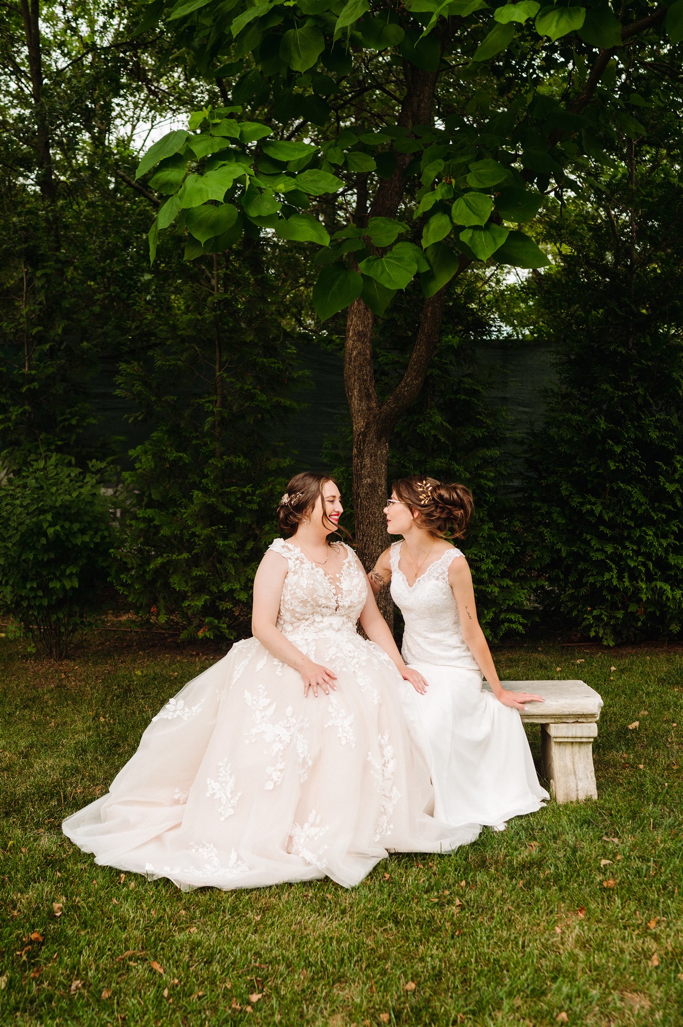 Two individuals in wedding dresses seated on a stone bench in a lush, green garden, holding hands.