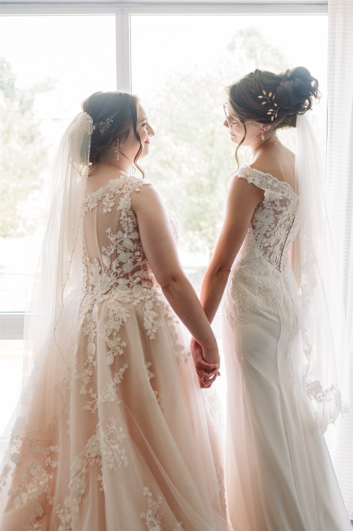 Two women in lace wedding dresses holding hands, facing each other in front of a bright window.