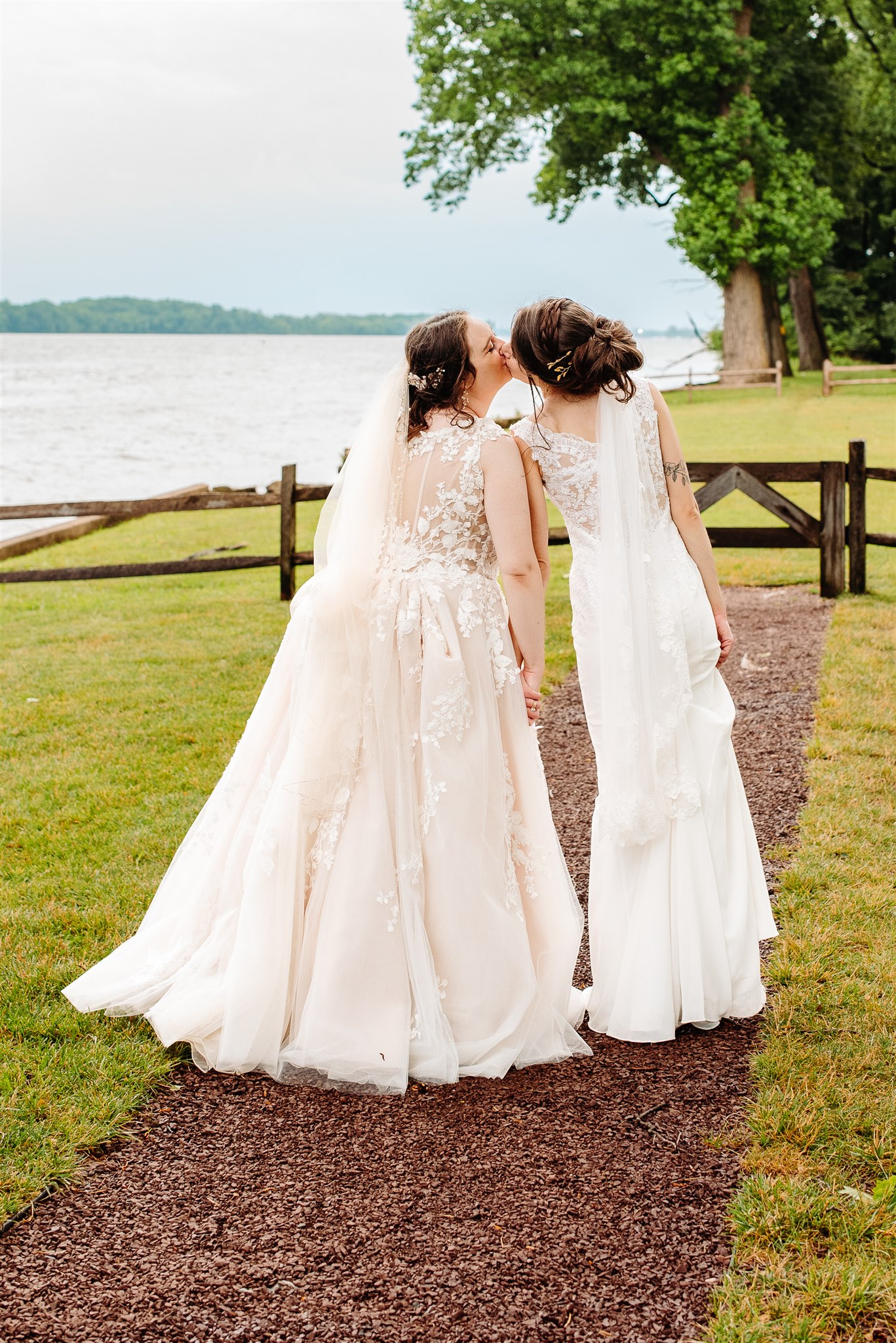 Two women in wedding gowns walk along a path by a lake, kissing with greenery and a fence behind them.