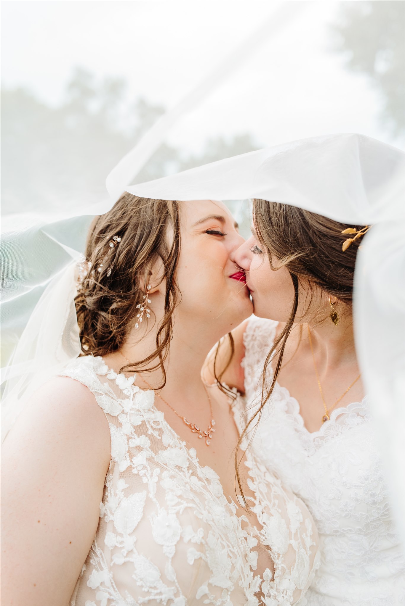 Two women kissing under a white veil, both in lace wedding dresses with jewelry, blurred background.