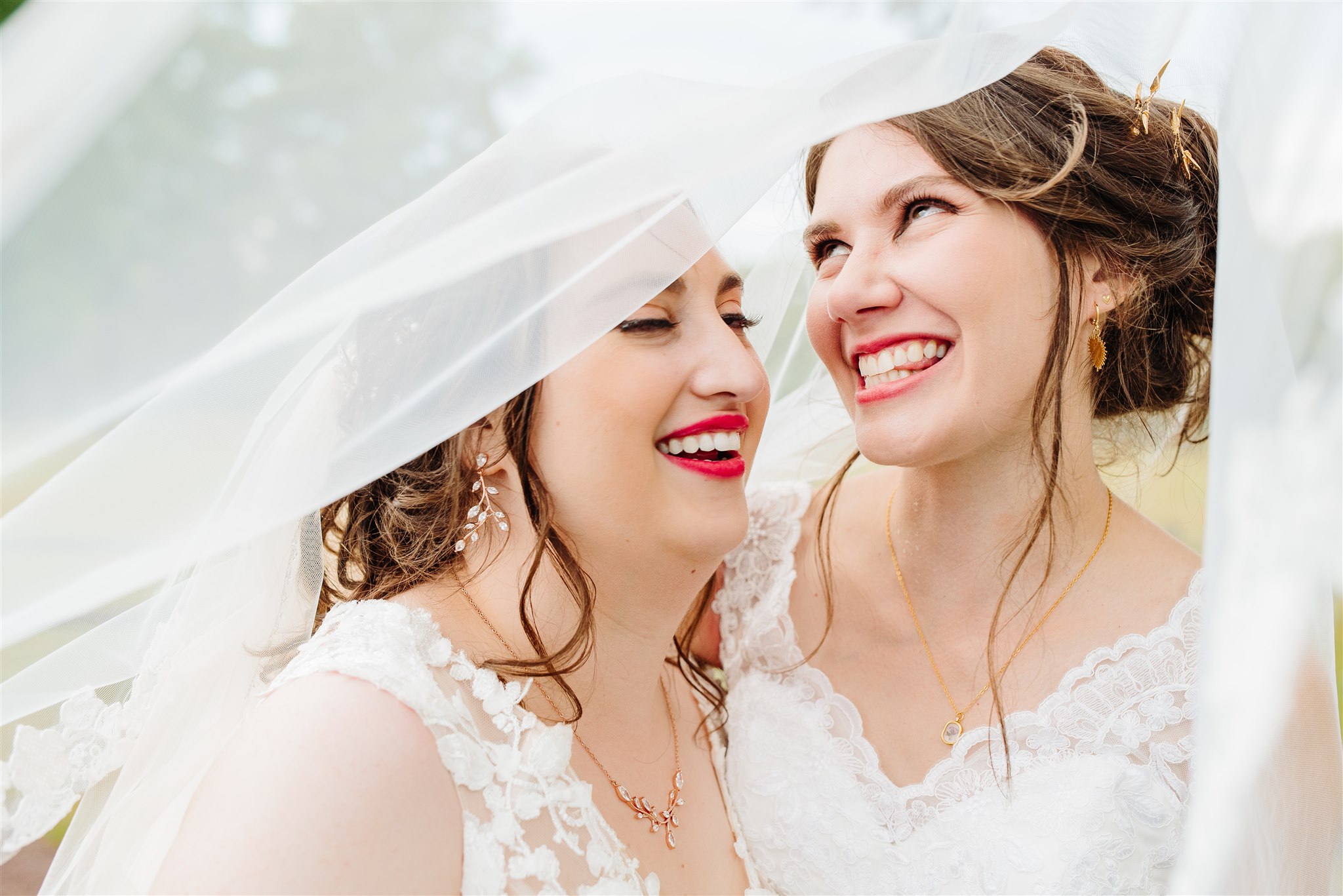 Two women in white lace dresses smiling, embracing, with flowing fabric and blurred background.