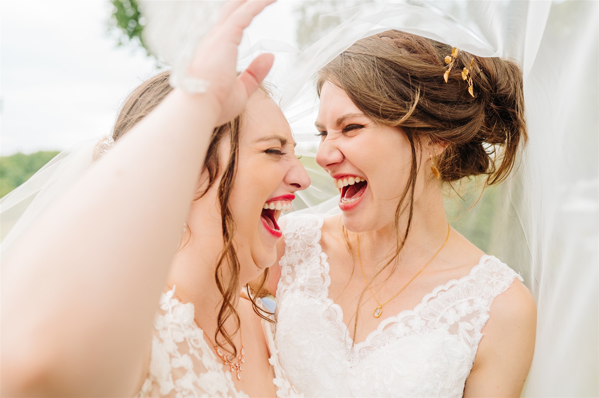 Two women in white lace dresses laugh joyfully under a light veil outdoors.