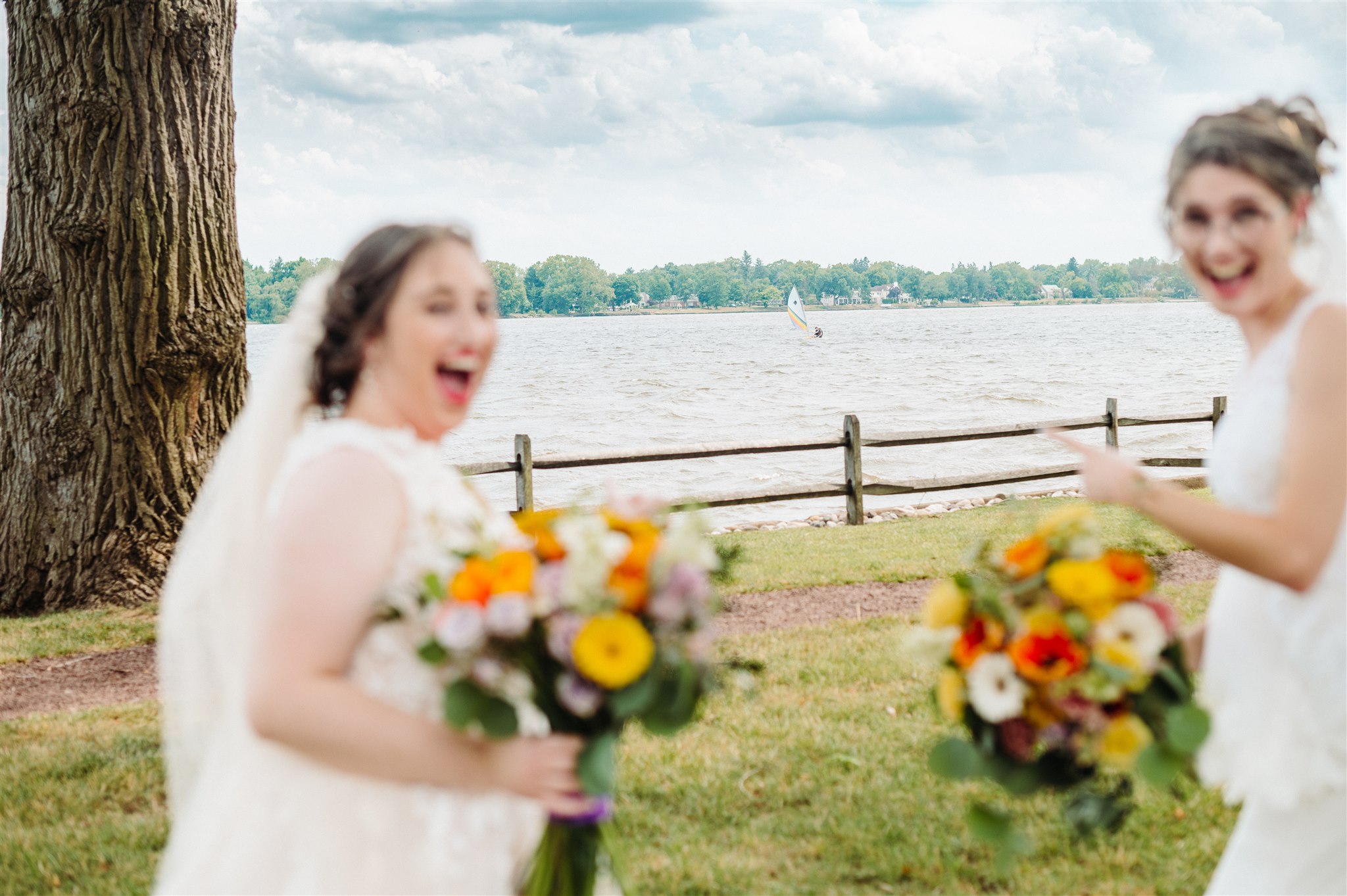 Two excited women in white dresses holding flower bouquets near a tree, overlooking water and a sailboat.