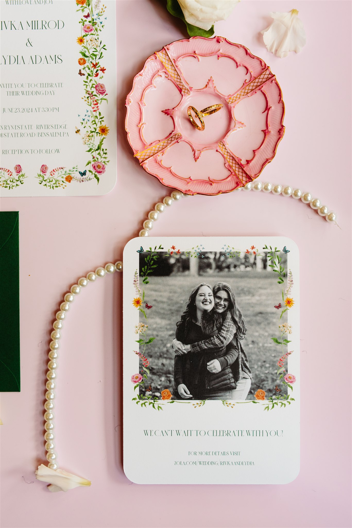 Wedding invitation suite on pink background with a floral card, couple photo, rings, and accessories.