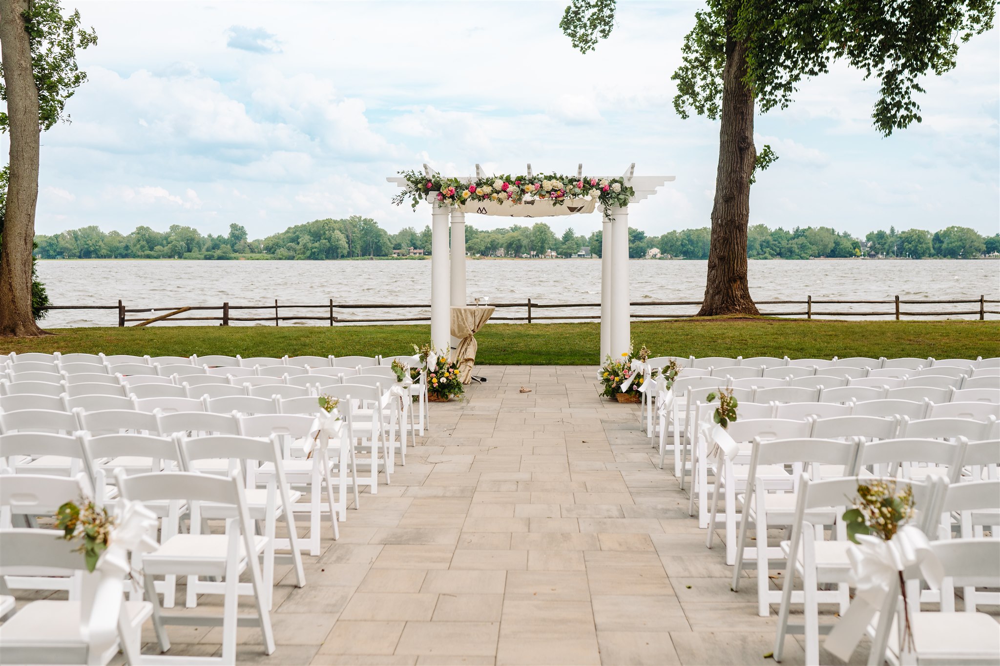 Outdoor wedding setup with white chairs and a flower-adorned arch by a lake with trees in view