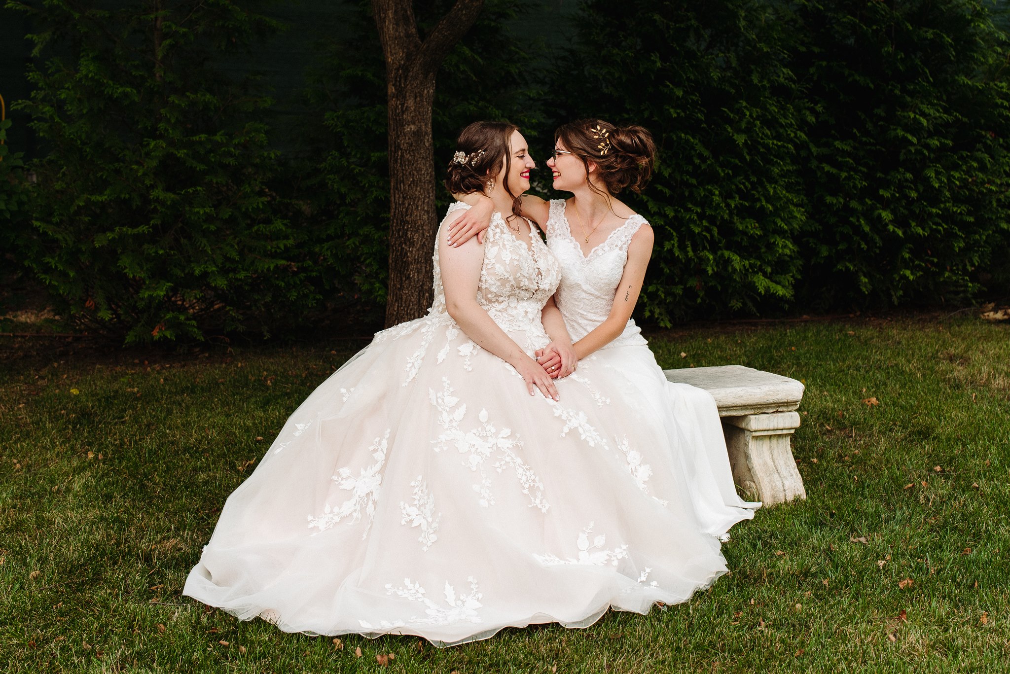 Two women in white lace wedding gowns sit closely on a stone bench in a lush outdoor setting.