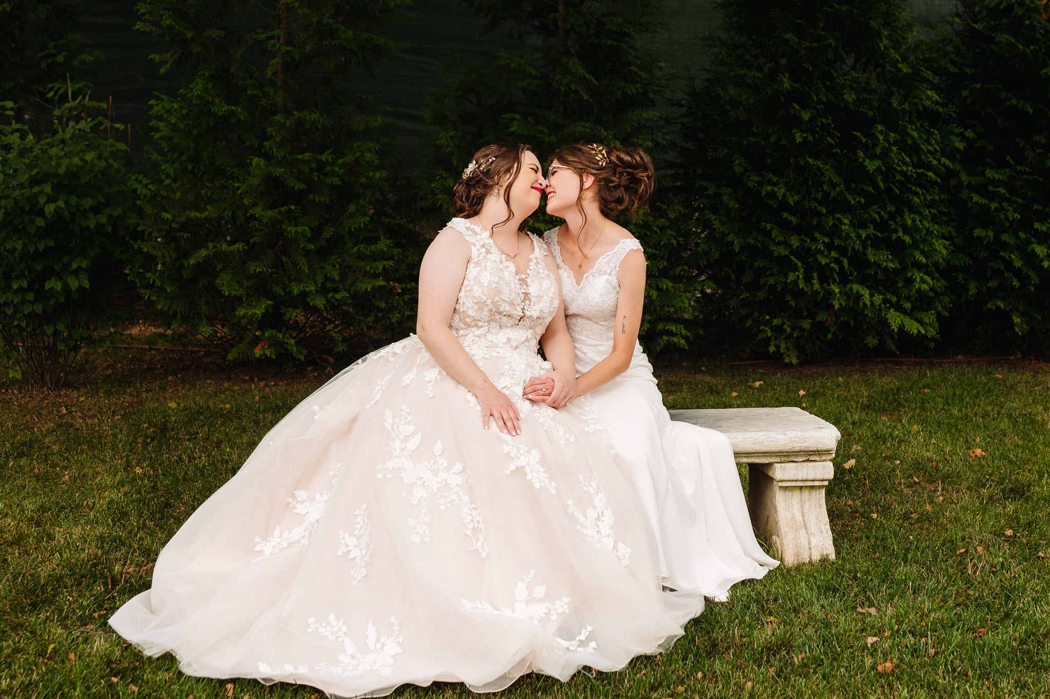 Two women in wedding gowns sit on a stone bench in a garden, holding hands and kissing.