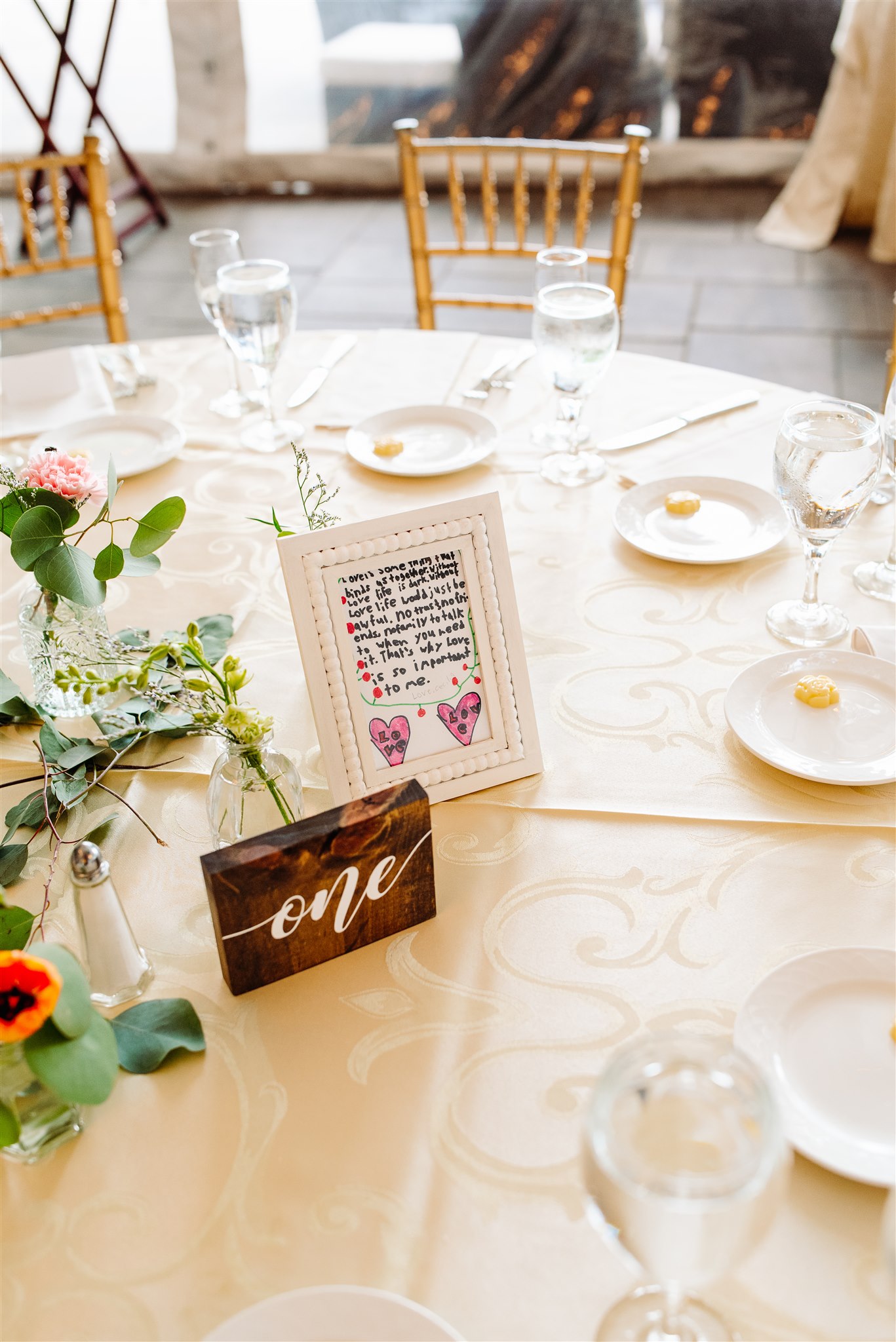 Wedding reception table with a cream tablecloth, gold chairs, floral arrangement, and a table number "one".