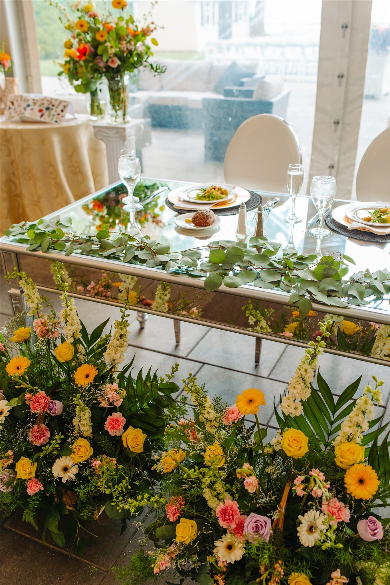 Glass-top dining table with green foliage, white chairs, place settings, and colorful floral arrangements.