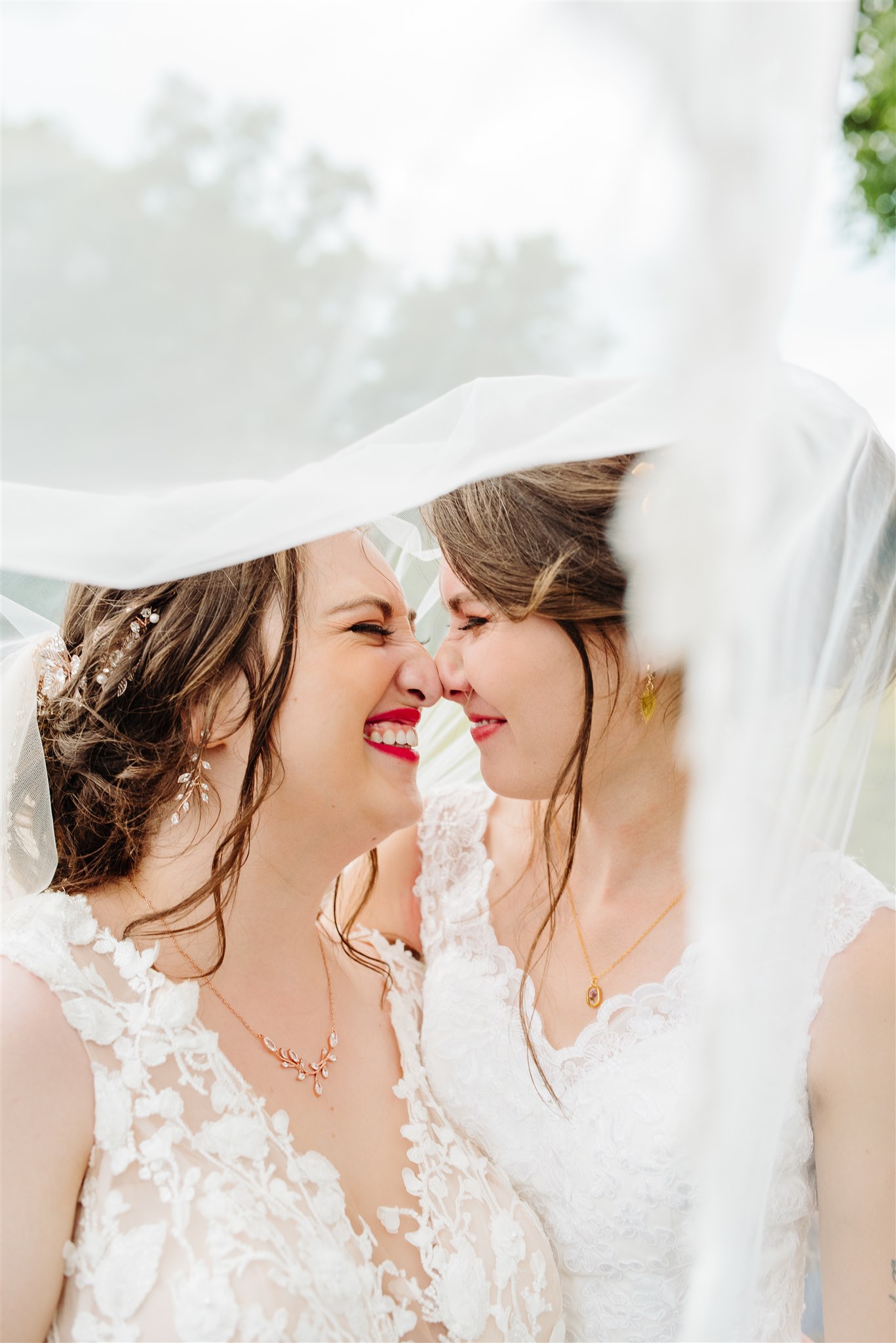 Two brides in white lace wedding gowns share an intimate, joyful moment under a veil.